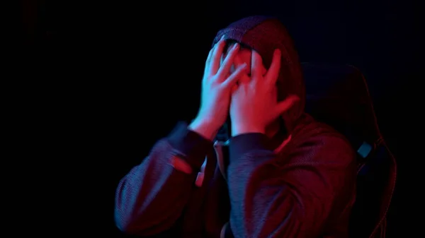 Ένας νεαρός άνδρας κάθεται σε έναν υπολογιστή και καλύπτει το πρόσωπό του με τα χέρια του. Ο άντρας με την κουκούλα κοιτάζει την οθόνη και ορκίζεται με θυμό. Προβολή πίσω από την οθόνη — Φωτογραφία Αρχείου