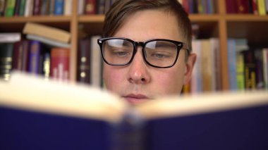 Genç bir adam kütüphanede kitap okuyor. Gözlüklü bir adam kitaba dikkatlice bakar. Arka planda kitap raflarında kitaplar var. Kitap kitaplığı.
