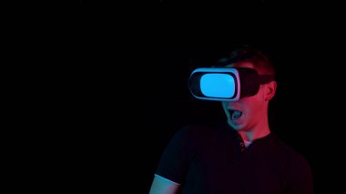 Young man in VR glasses. A man immersed in virtual reality makes movements with his hands. Blue and red light falls on a man on a black background.