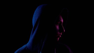 A young woman in a hood looks around. An attacker is standing in the dark. Blue and red light falls on a person on a black background.