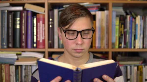 Молодий чоловік читає книжку в бібліотеці. Чоловік в окулярах уважно дивиться на книгу. На задньому плані знаходяться книги на книжкових полицях. бібліотека книг . — стокове фото