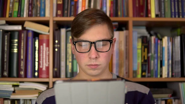 Genç bir adam tablette bir kitap okuyor. Gözlüklü bir adam tablete dikkatlice bakar. Arka planda kitap raflarında kitaplar var. Kitap kitaplığı. — Stok fotoğraf