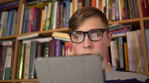 Молодий чоловік читає книжку в таблетці. Чоловік в окулярах уважно дивиться на планшет. На задньому плані знаходяться книги на книжкових полицях. бібліотека книг . — стокове фото