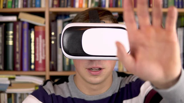 Anak muda berkacamata di perpustakaan. Seorang pria dengan helm VR di kepalanya memeriksa dan menyentuh realitas maya. Di latar belakangnya ada buku-buku di rak buku. Pustaka buku . — Stok Foto
