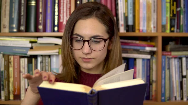 Молода жінка читає книжку в бібліотеці. Жінка в окулярах ретельно дивиться на книгу крупним планом. На задньому плані знаходяться книги на книжкових полицях. бібліотека книг . — стокове фото