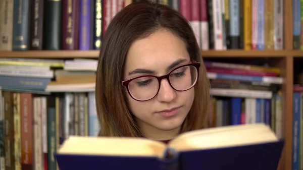 Mladá žena čte knihu v knihovně. Žena s brýlemi si pozorně prohlíží knihu. V pozadí jsou knihy na regálech. Knihovna knih. — Stock fotografie