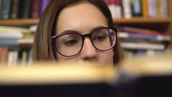 Μια νεαρή γυναίκα διαβάζει ένα βιβλίο σε μια βιβλιοθήκη. Μια γυναίκα με γυαλιά κοιτάζει προσεκτικά το βιβλίο από κοντά. Στο βάθος υπάρχουν βιβλία στα ράφια των βιβλιοθηκών. Βιβλιοθήκη βιβλίων. — Φωτογραφία Αρχείου