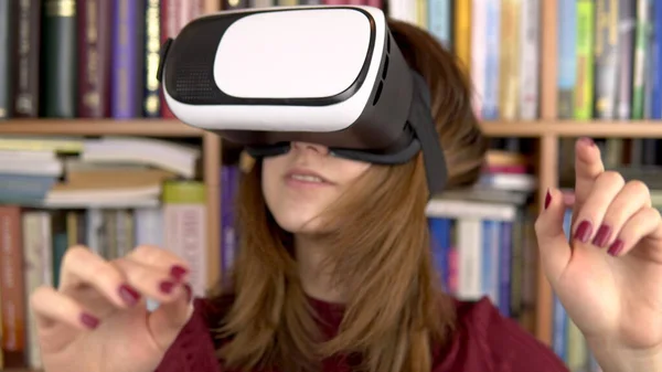 Молода жінка в окулярах VR в бібліотеці. Жінка з шоломом VR на голові вивчає і торкається віртуальної реальності. На задньому плані знаходяться книги на книжкових полицях. бібліотека книг . — стокове фото