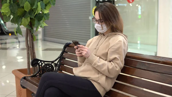 A young woman in a medical mask sits on a bench in a shopping center and talks on the phone. The masked woman protects herself from the epidemic of the Chinese virus "2019-nKoV". — Stock Photo, Image