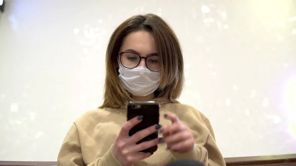 A young woman in a medical mask sits on a bench in a shopping center and talks on the phone. The masked woman protects herself from the epidemic of the Chinese virus 