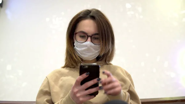 Uma jovem mulher em uma máscara médica senta-se em um banco em um centro comercial e fala ao telefone. A mulher mascarada protege-se da epidemia do vírus chinês "2019-nKoV ". — Fotografia de Stock