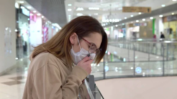 A young woman in a medical mask on the second floor in a shopping center. The woman is coughing. The masked woman protects himself from the epidemic of the Chinese virus "2019-nKoV". — Stock Photo, Image