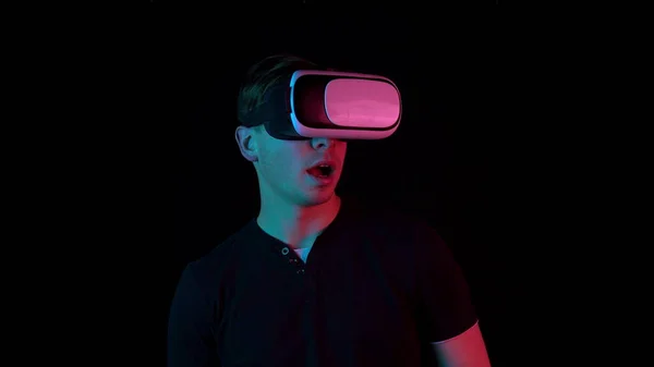 Młody człowiek w okularach Vr. Człowiek zanurzony w wirtualnej rzeczywistości wykonuje ruchy rękami. Niebieskie i czerwone światło pada na człowieka na czarnym tle. — Zdjęcie stockowe