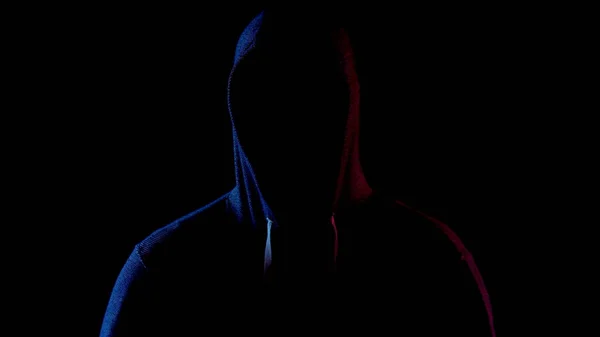 Un jeune homme se tient dans le capot et cache son visage. Un attaquant se tient dans le noir. La lumière bleue et rouge tombe sur la personne sur un fond noir . — Photo