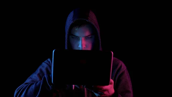 A young man in a hood with a laptop in his hands. A hacker makes a hack through a netbook. Blue and red light falls on a person on a black background.