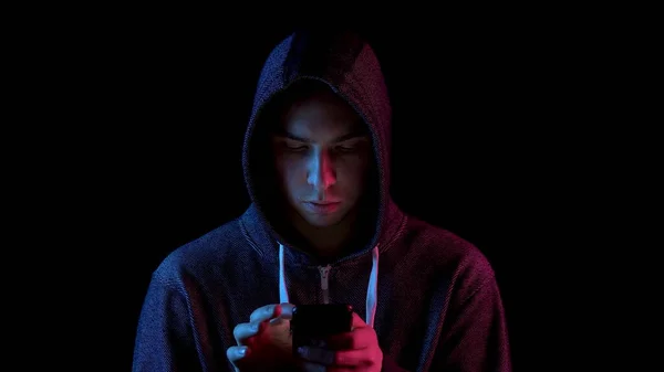 A young man in a hood with a phone in his hands. Hacker makes a hack through the phone. Blue and red light falls on a person on a black background.