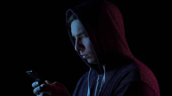Un ragazzo col cappuccio con un telefono in mano. L'hacker ha hackerato il telefono. Luce blu e rossa cade su una persona su uno sfondo nero . — Foto Stock