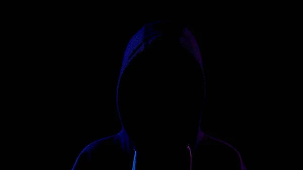 A young woman is standing in the hood hiding her face. An attacker is standing in the dark. Blue and red light falls on the person on a black background. — 스톡 사진