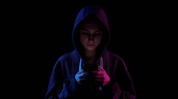 Una giovane donna in un cappuccio con un telefono in mano. L'hacker ha hackerato il telefono. Luce blu e rossa cade su una persona su uno sfondo nero . — Foto Stock