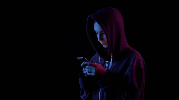 Una giovane donna in un cappuccio con un telefono in mano. L'hacker ha hackerato il telefono. Luce blu e rossa cade su una persona su uno sfondo nero . — Foto Stock