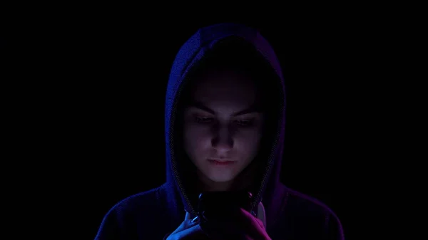 A young woman in a hood with a phone in her hands closeup. Hacker makes a hack through the phone. Blue and red light falls on a person on a black background. — Stockfoto