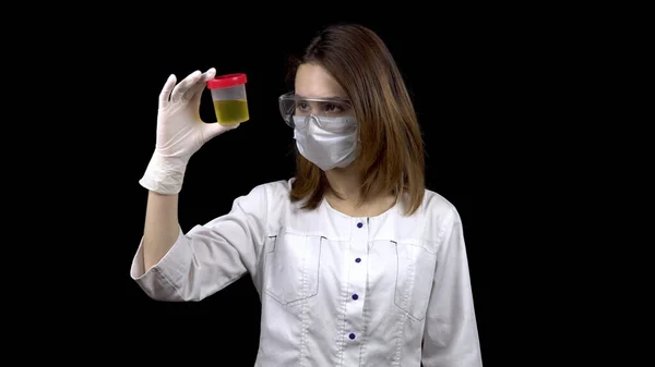 Una giovane dottoressa sta controllando gli esami delle urine. Una donna esamina un vaso di prova contenente urina su uno sfondo nero . — Foto Stock