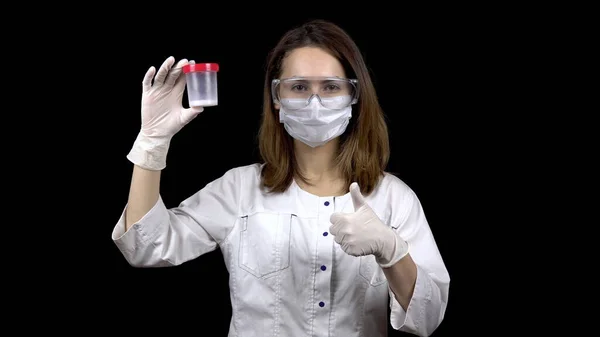 Jeune femme médecin vérifie les tests de sperme. Une femme examine un bocal de test, puis montre un pouce vers le haut. Analyse positive du sperme. Sur fond noir . — Photo
