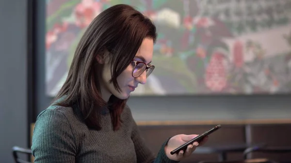 Een jonge vrouw zit in een café met een telefoon. Het meisje is in correspondentie op een smartphone. Close-up. — Stockfoto