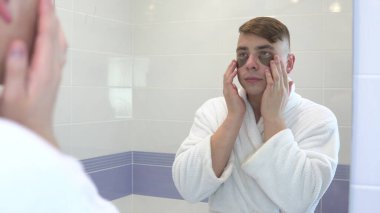 Genç bir adam yüzüne kozmetik bantlar yapıştırıyor. Spa tedavileri. Deri yenilenmesi için kahverengi hidrojel yamalar. Banyodaki aynanın önünde beyaz bir bornozla duruyor. Aynaya bak.