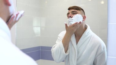 Genç bir adam aynanın önünde tıraş köpüğü sürer. Yüzünde köpük olan beyaz önlüklü bir adam. Aynaya bak..