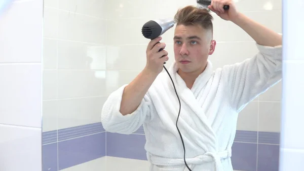 Um jovem seca o cabelo com um secador de cabelo. Um homem de casaco branco faz styling usando um secador de cabelo e um pente. Fica em frente a um espelho do banheiro. Vista através do espelho — Fotografia de Stock