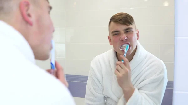 Un joven se cepilla los dientes activamente. Un hombre con un albornoz blanco frente a un espejo de baño. Vista a través del espejo —  Fotos de Stock