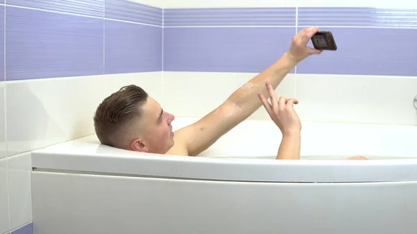 Молодой человек лежит в ванной и делает селфи по телефону. Мужчина расслабляется в ванной . — стоковое фото