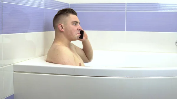Um jovem está deitado no banho e a falar ao telefone. Um homem relaxa no banho — Fotografia de Stock