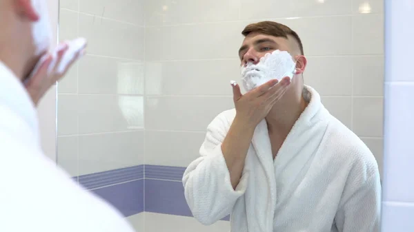 Un joven aplica espuma de afeitar delante de un espejo. Un hombre con un abrigo blanco con espuma en la cara. Vista a través del espejo . —  Fotos de Stock