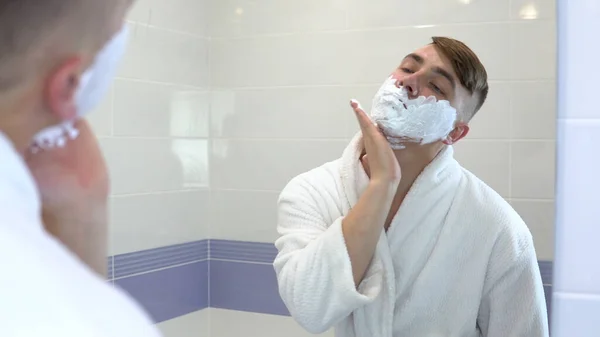 Un joven aplica espuma de afeitar delante de un espejo. Un hombre con un abrigo blanco con espuma en la cara. Vista a través del espejo . —  Fotos de Stock
