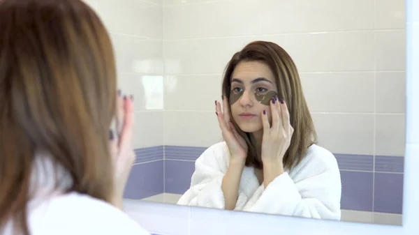 Young woman puts cosmetic patches on her face. Spa treatments. Brown hydrogel patches for skin rejuvenation. Woman is standing in a white bathrobe in front of the bathroom mirror. View through the