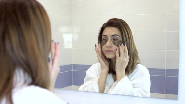 La joven se pone parches cosméticos en la cara. Tratamientos de spa. Parches de hidrogel marrón para el rejuvenecimiento de la piel. La mujer está de pie en un albornoz blanco frente al espejo del baño. Vista a través de la —  Fotos de Stock