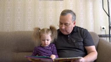 Torunu büyükbabasıyla kitap okuyor. Kız kitaba kaşlarını çatıyor ve büyükbabayı dikkatle dinliyor. Kanepede birlikte oturuyoruz. Yakın çekim.