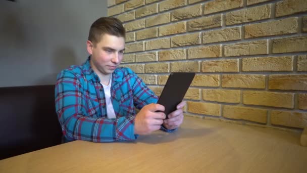 Um jovem com uma tábua senta-se à mesa. Um homem senta-se em casa em uma mesa com um tablet e está em correspondência . — Vídeo de Stock