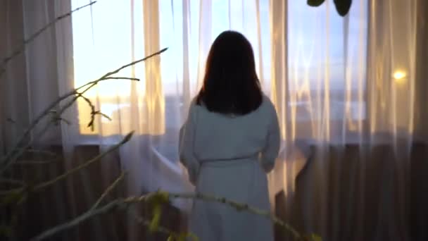 Beyaz önlüklü genç bir kadın pencereye doğru yürür ve perdeleri açar. Sabah kız pencereden dışarı bakar ve parlak güneş parlar.. — Stok video