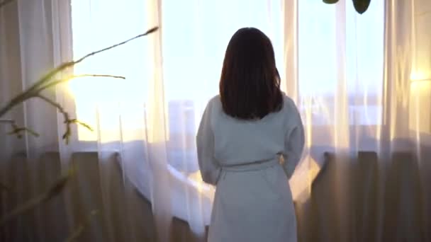 A young woman in a white coat walks to the window and opens the curtains. The girl in the morning looks out the window and the bright sun shines. — Stock Video