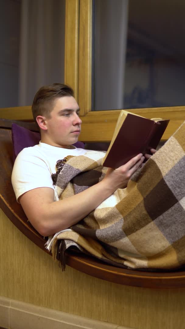 一个年轻人在看书。一个人躺在窗台上，手里拿着一本书。到了晚上。垂直录像. — 图库视频影像