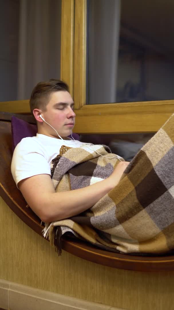 Un joven escucha música en los auriculares. Un hombre yace en un alféizar junto a la ventana con auriculares en los oídos. Afuera de la noche. Vídeo vertical . — Vídeos de Stock