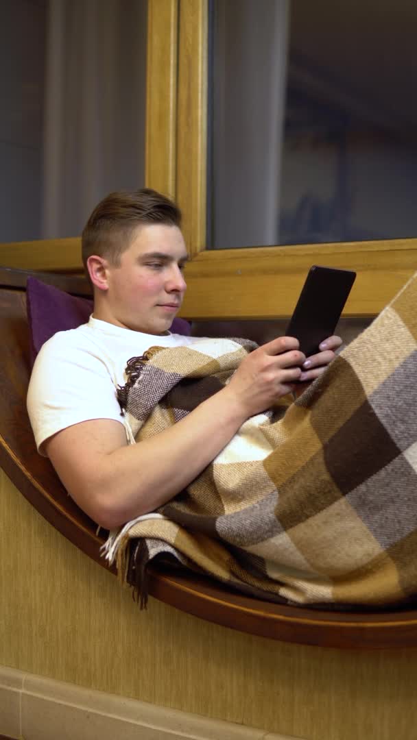 Молодой человек пишет смс на планшете. Мужчина лежит на подоконнике у окна с табличкой в руках. За вечерним окном. Вертикальное видео . — стоковое видео