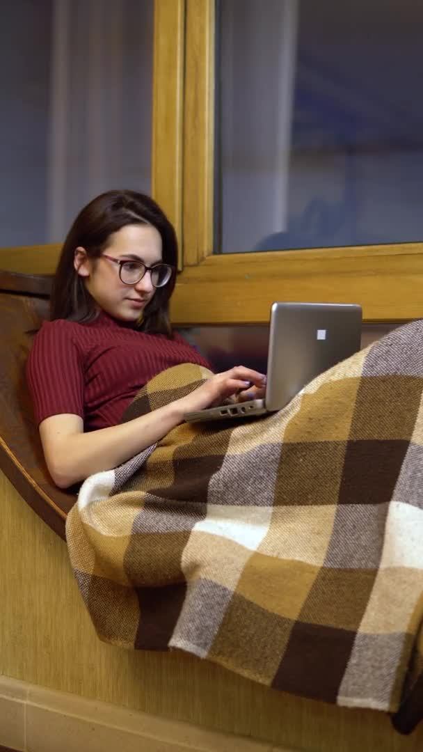 Молодая женщина печатает на ноутбуке. Девушка лежит на подоконнике у окна с ноутбуком в руках. На улице вечером. Вертикальное видео . — стоковое видео