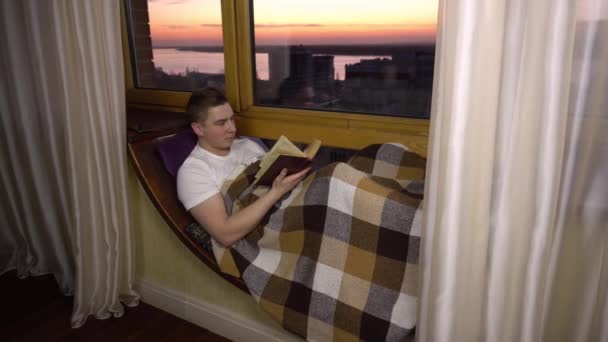 Un joven está leyendo un libro. Un hombre yace en un alféizar junto a la ventana con un libro en sus manos. Fuera de la noche — Vídeos de Stock