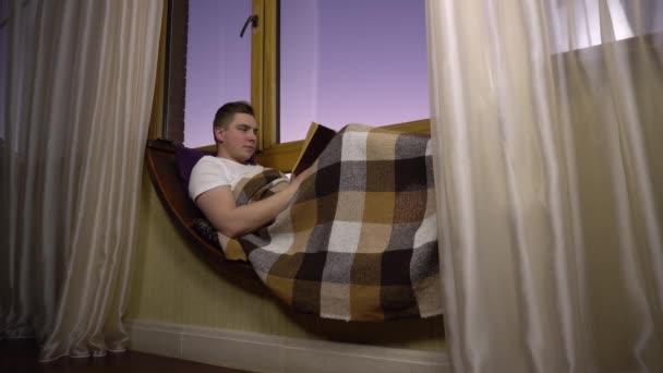 Ein junger Mann liest ein Buch. Ein Mann liegt mit einem Buch in der Hand auf einem Fensterbrett am Fenster. Aus dem Abend — Stockvideo