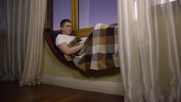 Een jongeman luistert naar muziek op een koptelefoon. Een man ligt op een vensterbank bij het raam met een koptelefoon in zijn oren. Buiten de avond. — Stockvideo