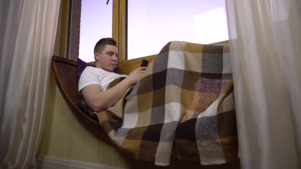 Un joven está enviando mensajes por teléfono. Un hombre yace en un alféizar junto a la ventana con un teléfono inteligente en sus manos. Fuera de la ventana de la noche . — Vídeos de Stock